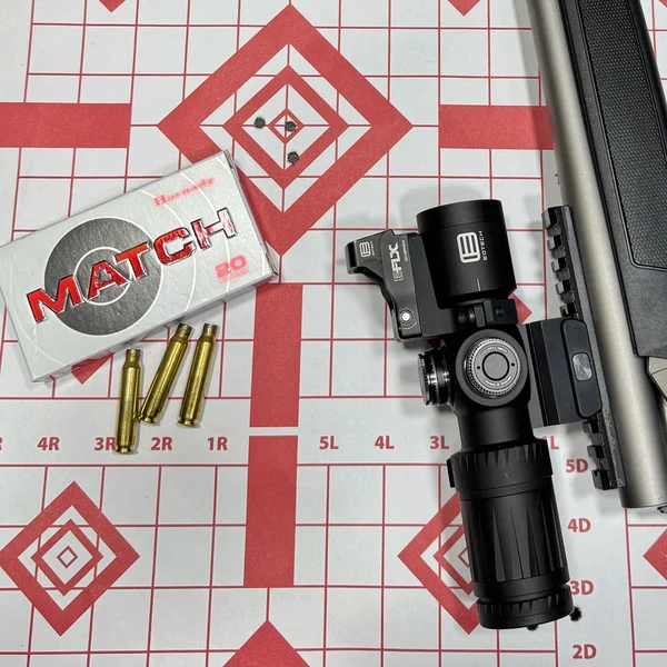 Three-shot accuracy group on a red diamond target next to Hornady Match .223 ammunition and a rifle scope
