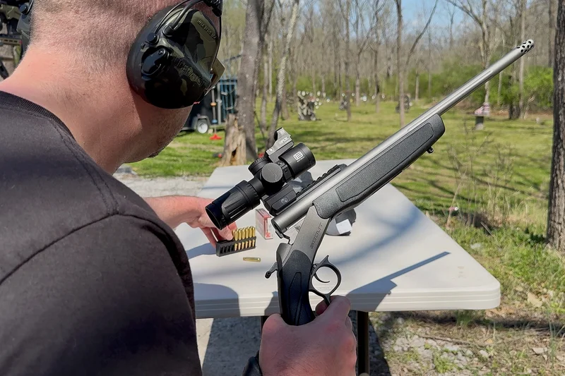 Shooter using a CVA Scout TD rifle with a mounted scope at an outdoor shooting range bench