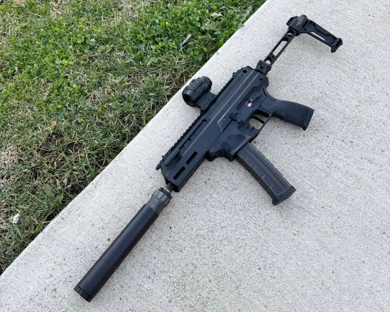 Compact firearm equipped with Silencer Central BANISH 45 suppressor resting on concrete beside grass