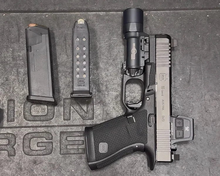 Overhead flat lay of a Glock 19 handgun equipped with a Vortex optic, a SureFire weapon light, and two loaded magazines