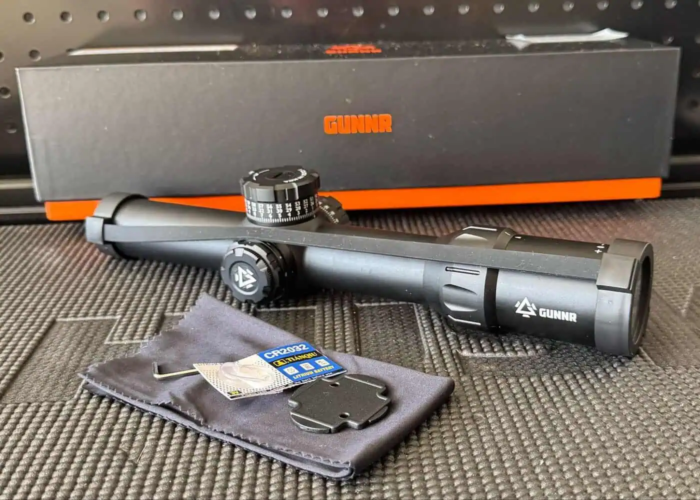 GUNNR Odin LPVO rifle optic with included accessories on a table, including throw lever, torque key, CR2032 battery, turret cap tool, lens covers, and lens cloth, with branded box in background