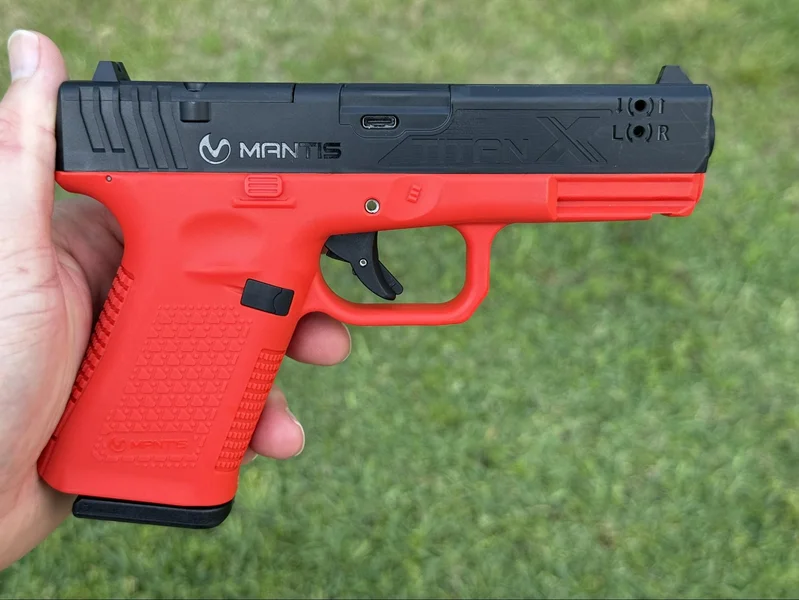 The Mantis TitanX laser training pistol held in a hand, showcasing the high-visibility red frame and black slide used for safe dry-fire practice